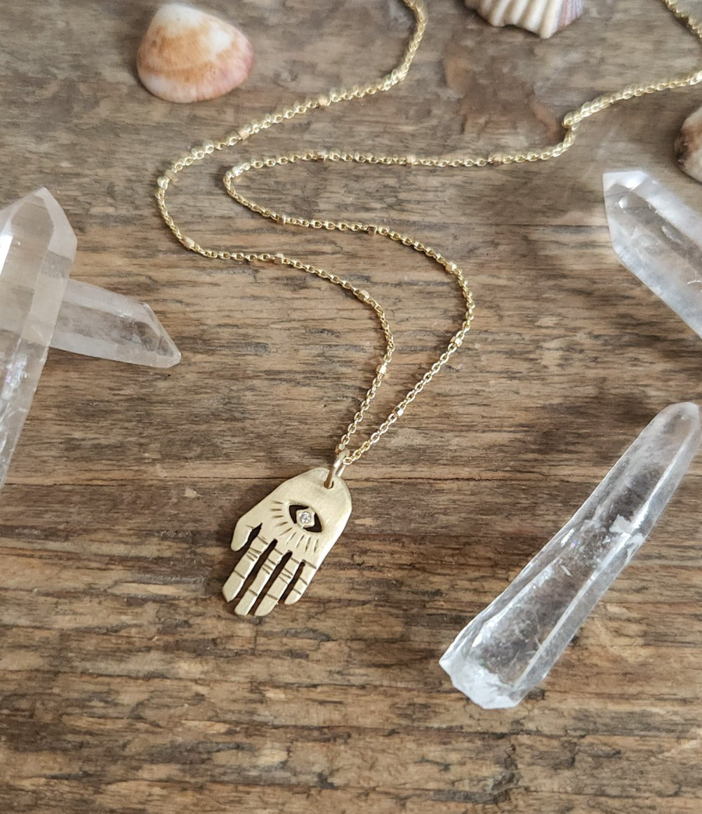 A golden necklace with a white diamond pendant in the shape of Hamsa hand