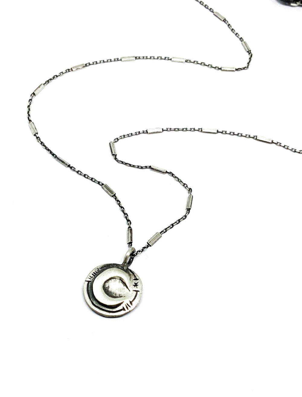 A silver necklace with a pendant featuring a stylized sun and moon design, on a white background.