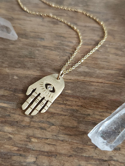 A golden necklace with a white diamond pendant, featuring a brushed finish and a Hamsa hand pendant. The necklace is displayed on wooden background