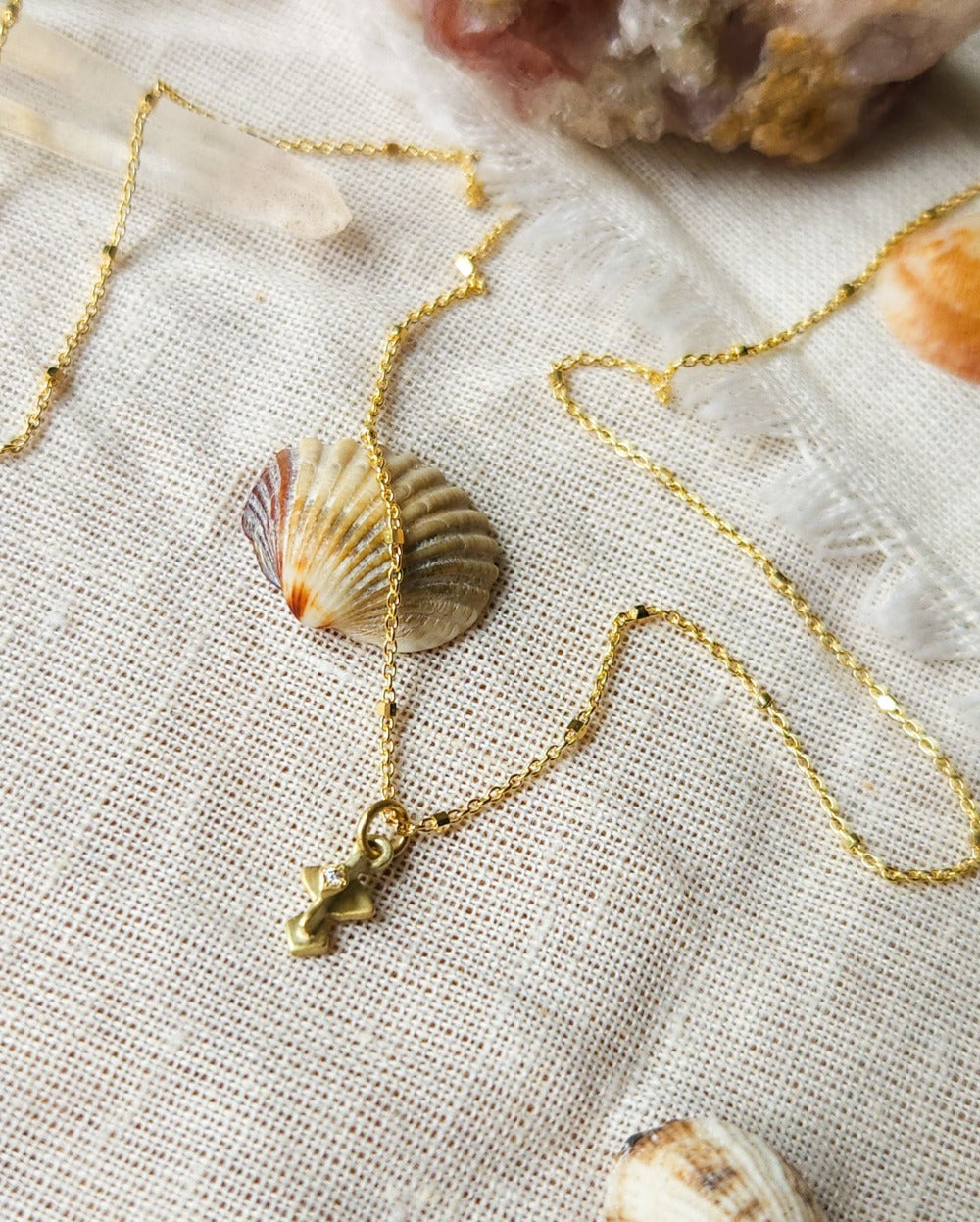 A golden necklace with a white diamond pendant, featuring a brushed finish and a Hamsa talisman pendant. The necklace is displayed on a textured fabric with decorative stones and crystals.
