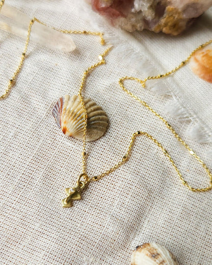 A golden necklace with a white diamond pendant, featuring a brushed finish and a Hamsa talisman pendant. The necklace is displayed on a textured fabric with decorative stones and crystals.