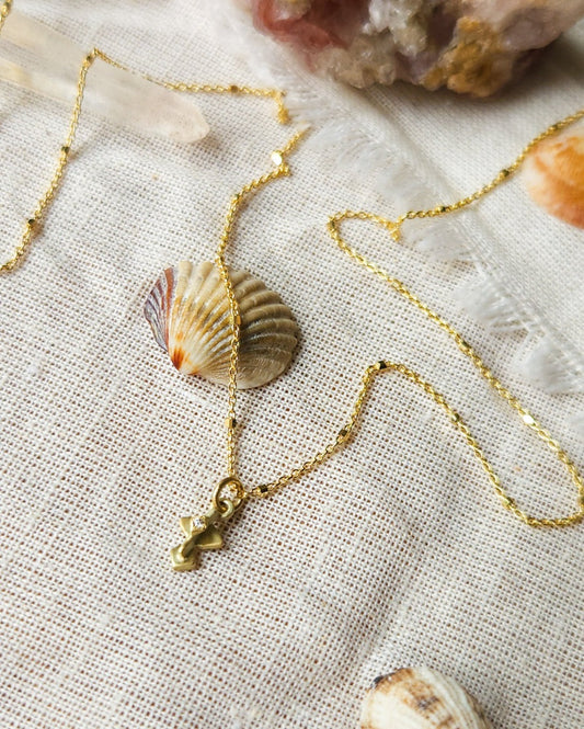 A golden necklace with a white diamond pendant, featuring a brushed finish and a Hamsa talisman pendant. The necklace is displayed on a textured fabric with decorative stones and crystals.