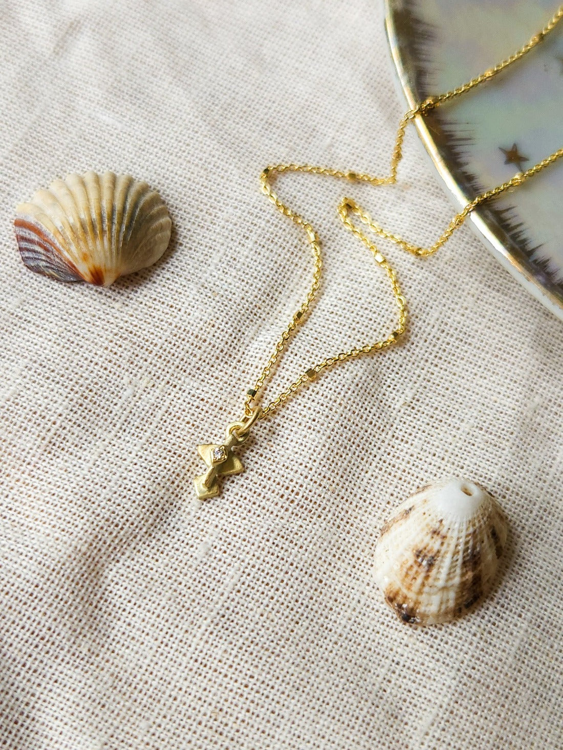 A golden necklace with a white diamond pendant, featuring a brushed finish and a Hamsa talisman pendant. The necklace is displayed on a textured fabric with decorative stones and crystals.