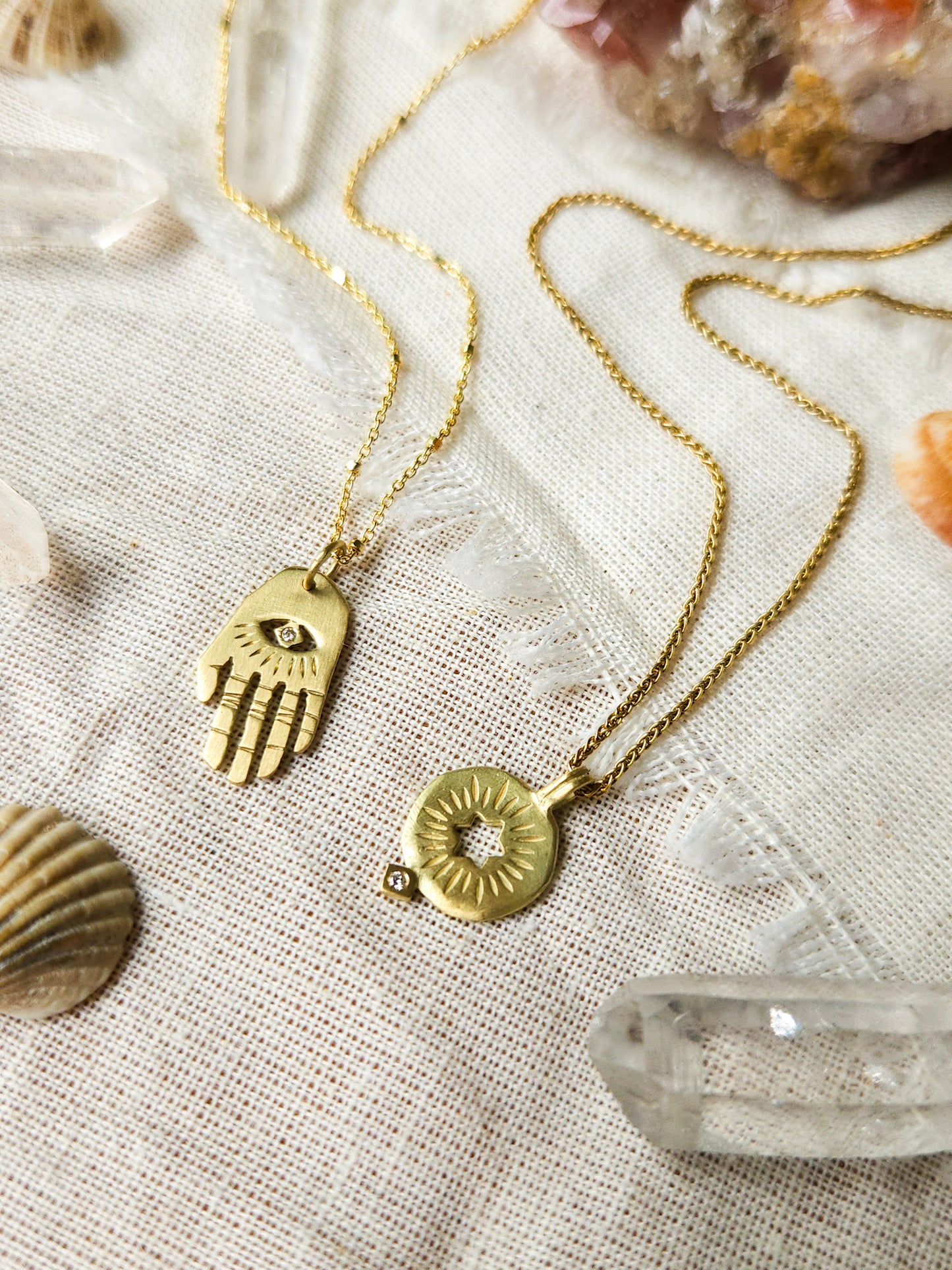 A golden necklace with a white diamond pendant in the shape of Hamsa hand next to pendant with a cut out rising star of david