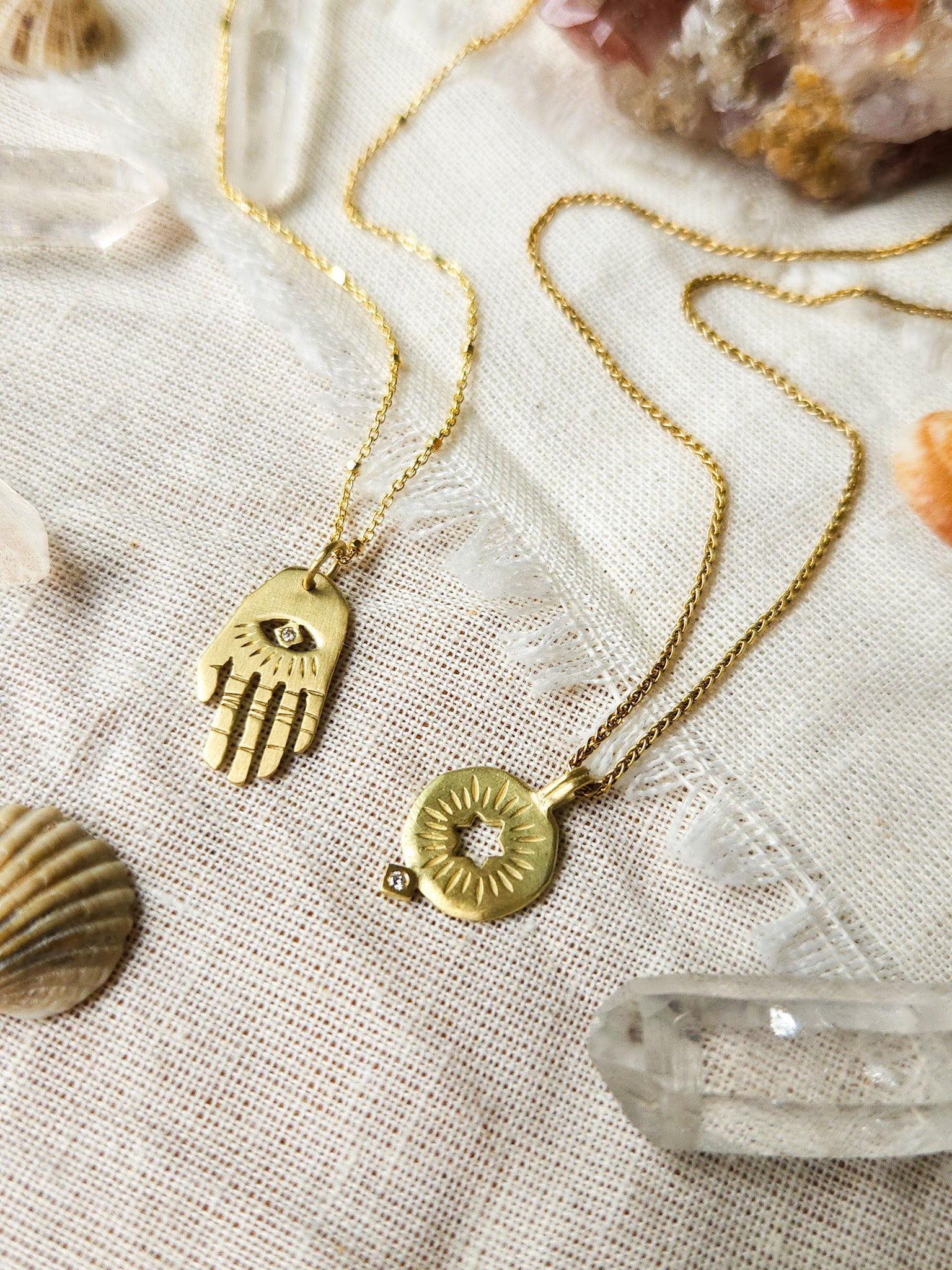 A golden necklace with a white diamond pendant in the shape of Hamsa hand next to pendant with a cut out rising star of david