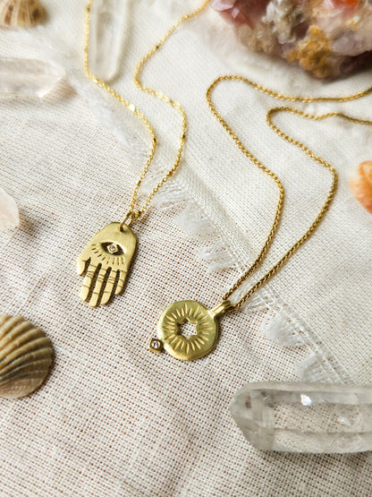 A golden necklace with a white diamond pendant in the shape of Hamsa hand next to pendant with a cut out rising star of david