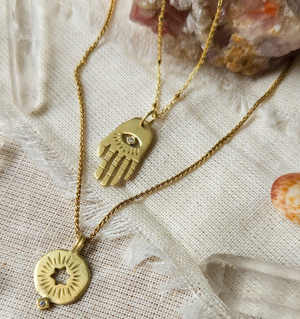 A golden necklace with a white diamond pendant in the shape of Hamsa hand next to pendant with a cut out rising star of david