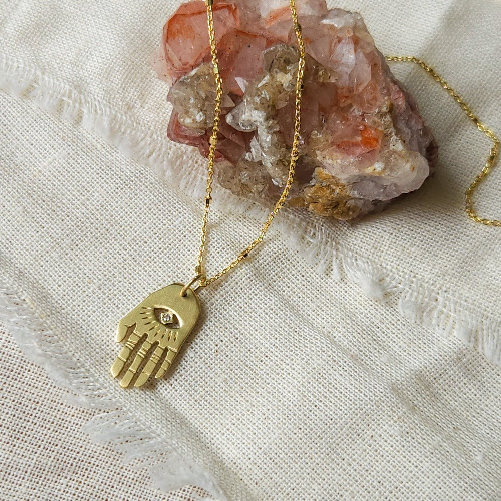 A golden Hamsa necklace with a white diamond pendant, displayed on a textured background.