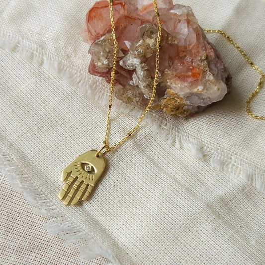A golden Hamsa necklace with a white diamond pendant, displayed on a textured background.