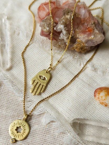 A golden necklace with a white diamond pendant in the shape of Hamsa hand next to pendant with a cut out rising star of david