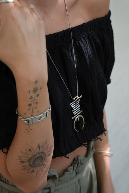 A silver and brass necklace with a geometric pendant, featuring a chain with small bell-like charms and a larger brass ring pendant.