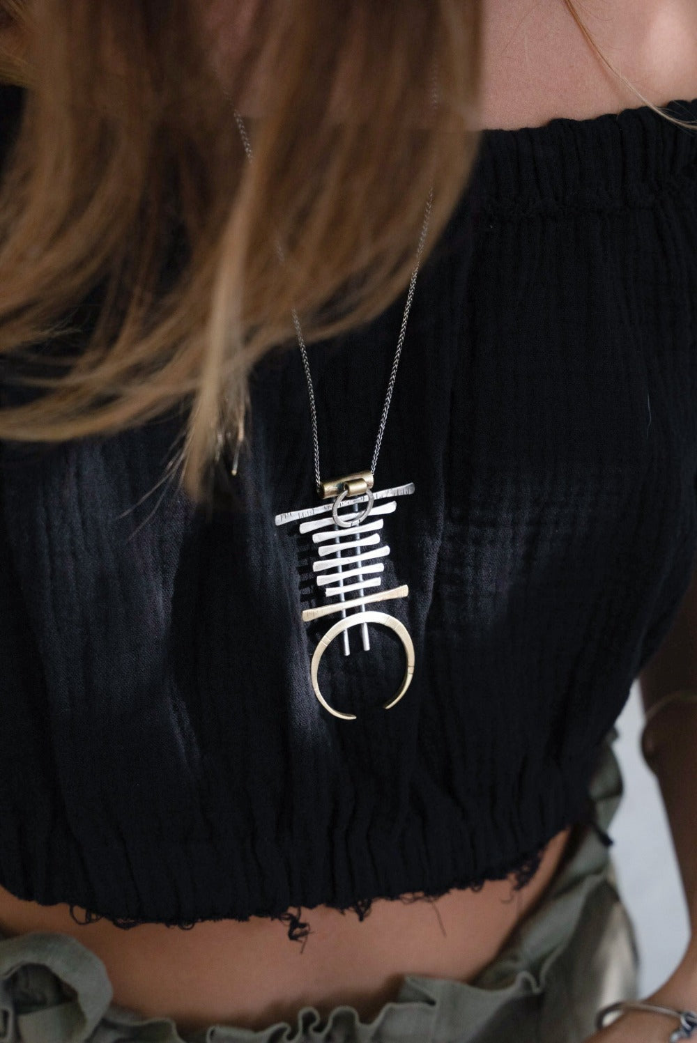 A silver and brass necklace with a geometric pendant, featuring a chain with small bell-like charms and a larger brass ring pendant.