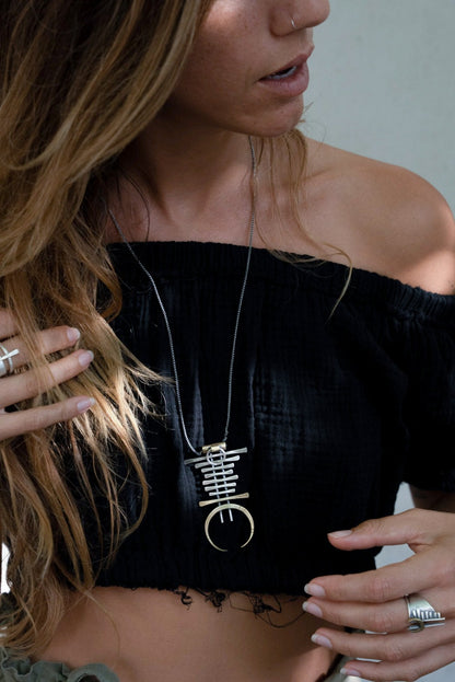 A silver and brass necklace with a geometric pendant, featuring a chain with small bell-like charms and a larger brass ring pendant.