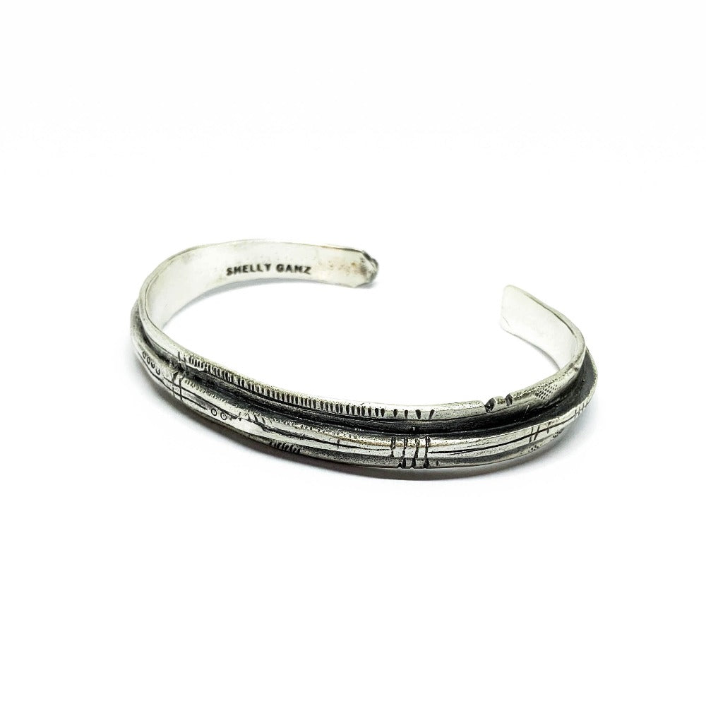 A sterling silver bracelet with an oxidized and brushed finish, featuring text on the bracelet surface.