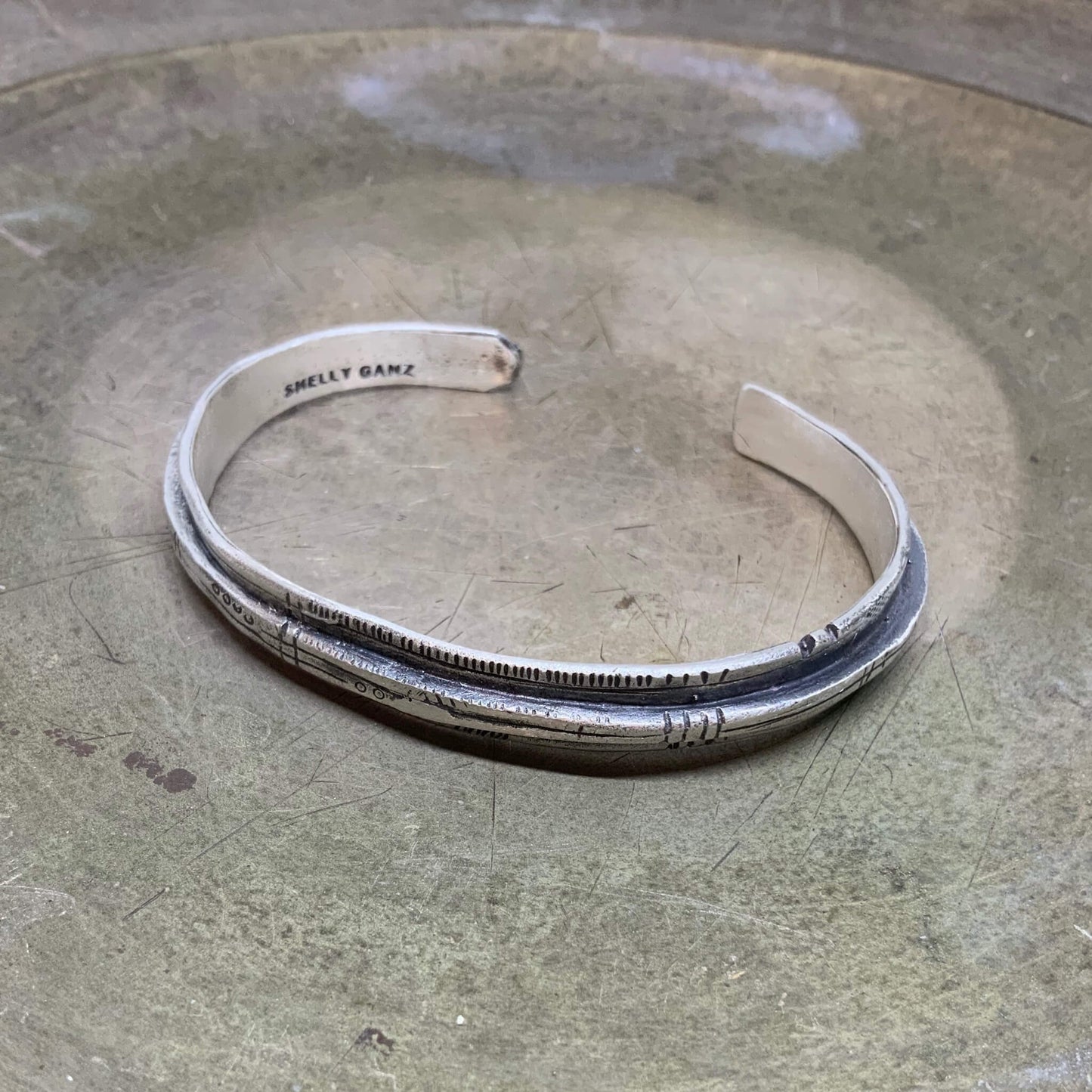 A sterling silver bracelet with an oxidized and brushed finish, featuring text on the bracelet surface.