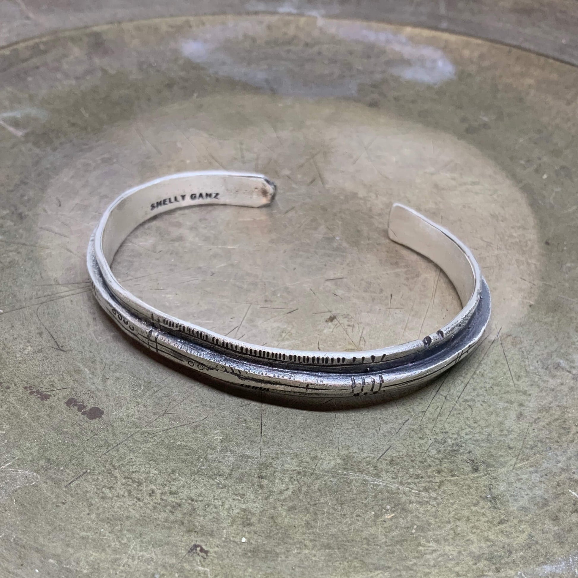 A sterling silver bracelet with an oxidized and brushed finish, featuring text on the bracelet surface.