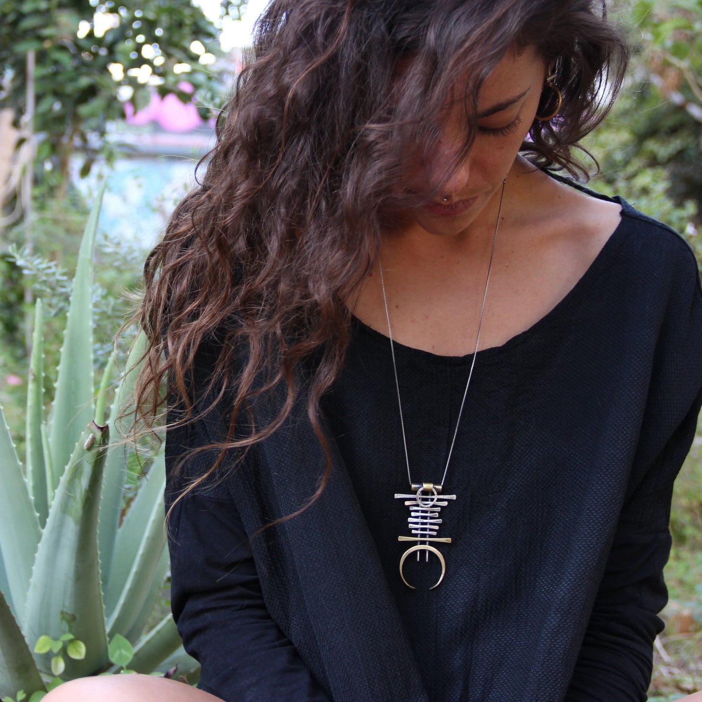 A silver and brass necklace with a geometric pendant, featuring a chain with small bell-like charms and a larger brass ring pendant.