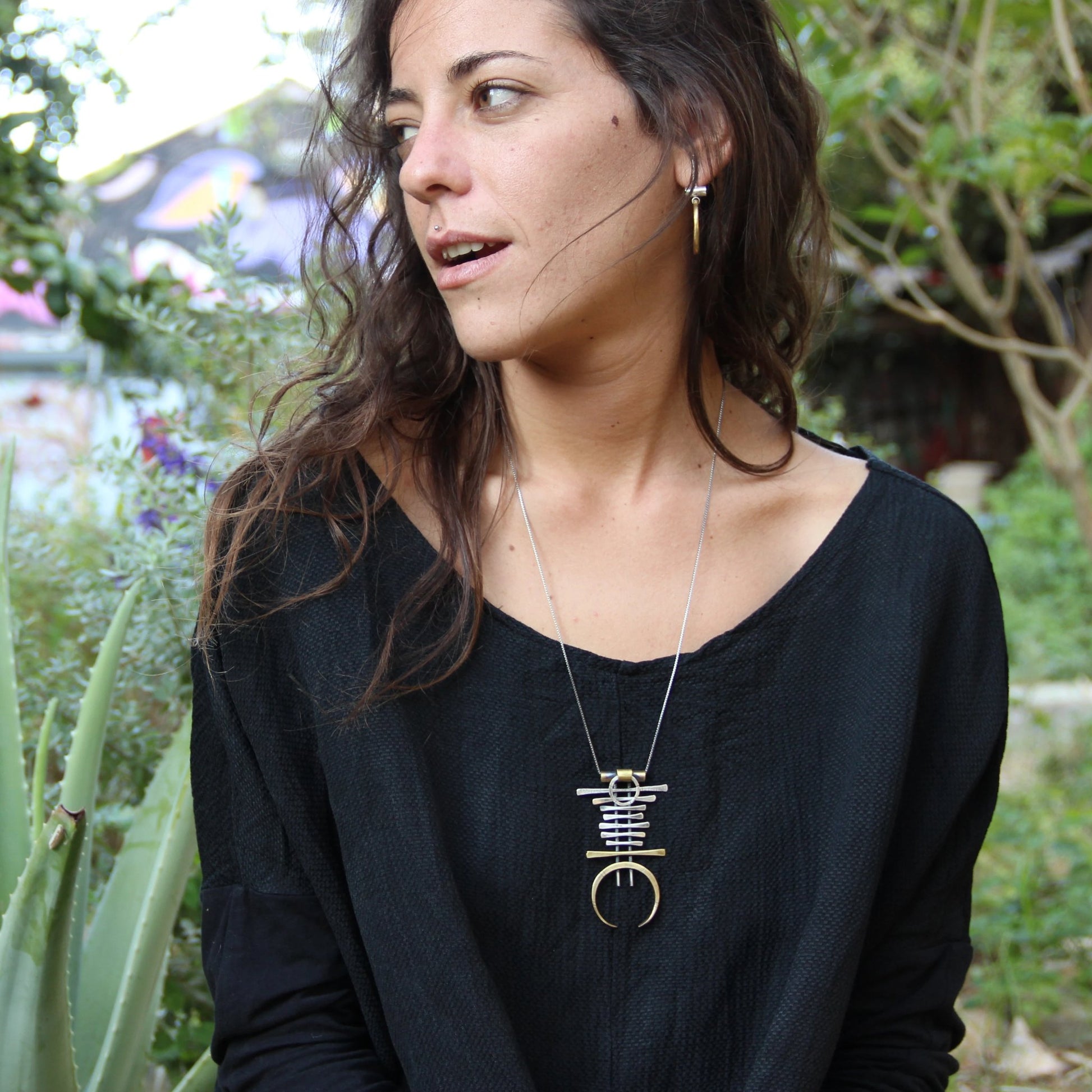 A silver and brass necklace with a geometric pendant, featuring a chain with small bell-like charms and a larger brass ring pendant.