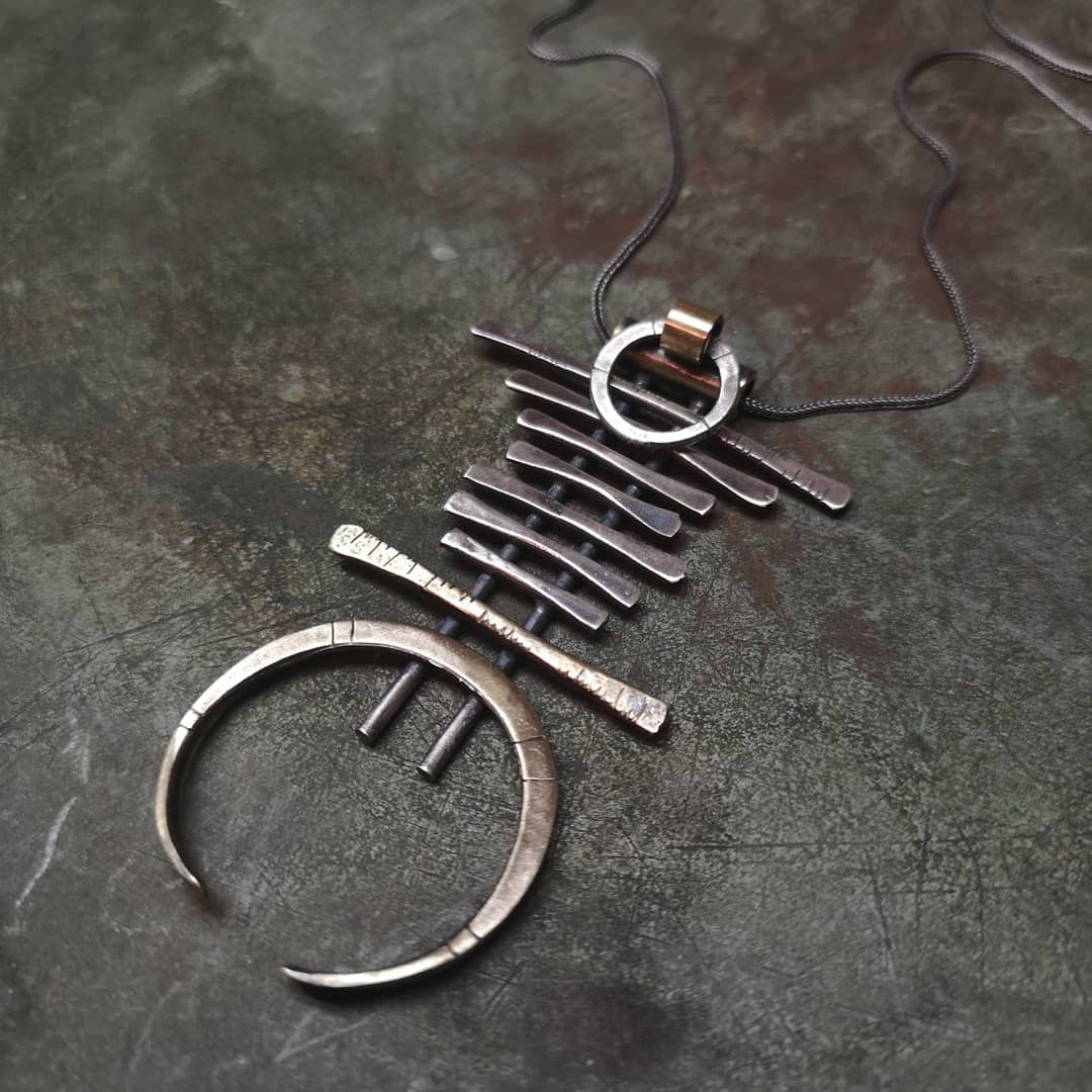 A silver and brass necklace with a geometric pendant, featuring a chain with small bell-like charms and a larger brass ring pendant.
