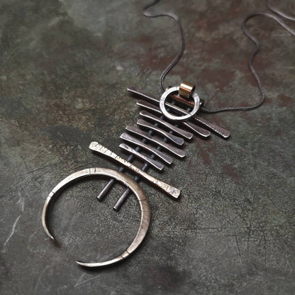 A silver and brass necklace with a geometric pendant, featuring a chain with small bell-like charms and a larger brass ring pendant.