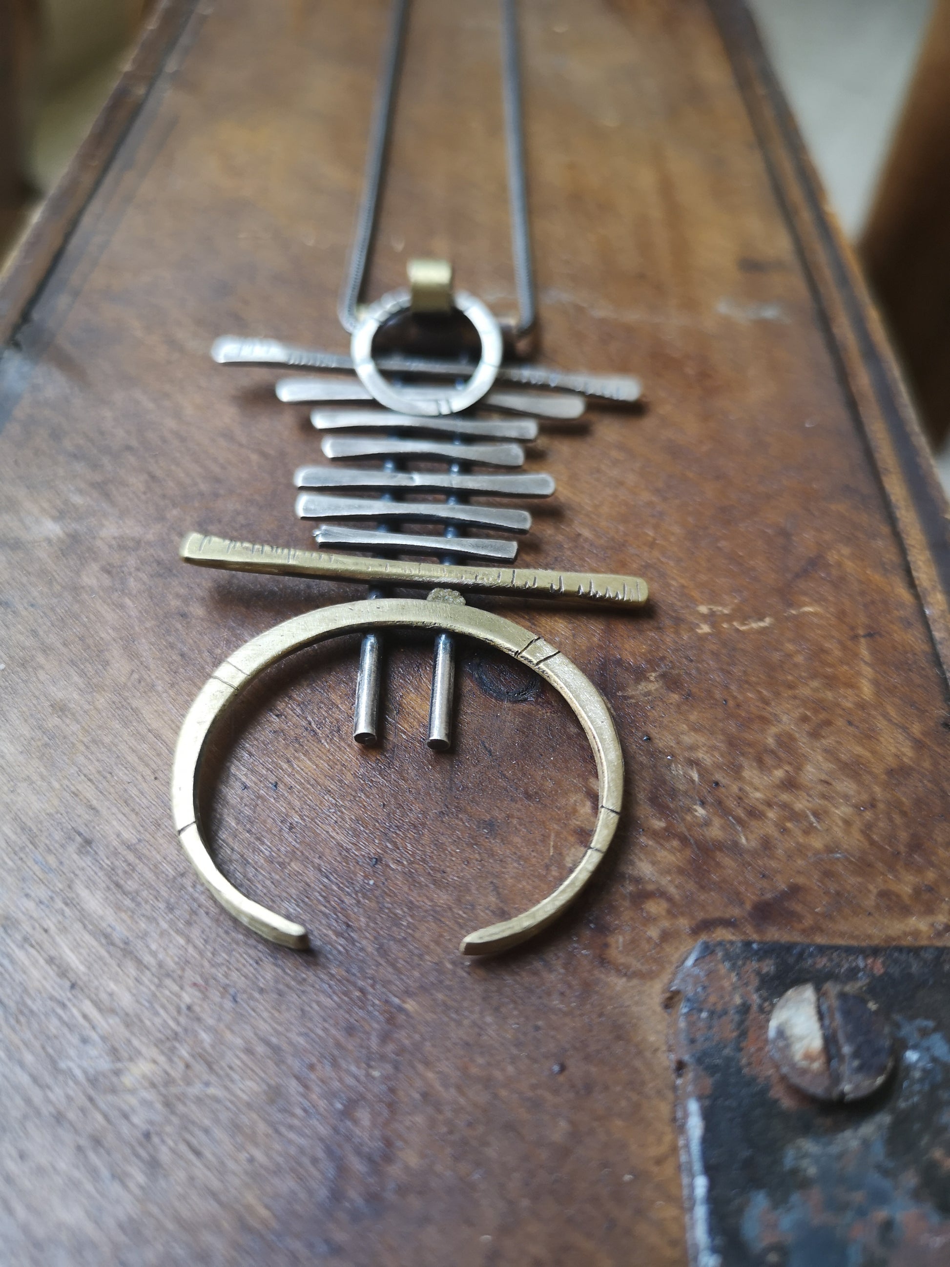 A silver and brass necklace with a geometric pendant, featuring a chain with small bell-like charms and a larger brass ring pendant.