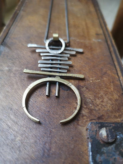 A silver and brass necklace with a geometric pendant, featuring a chain with small bell-like charms and a larger brass ring pendant.