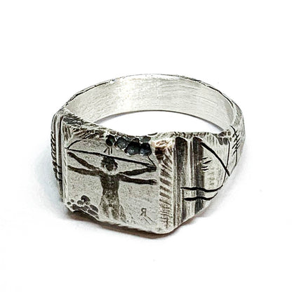 A silver ring with an oxidized and brushed finish, featuring an engraved design inspired by Florentine art, including figures and architectural elements.