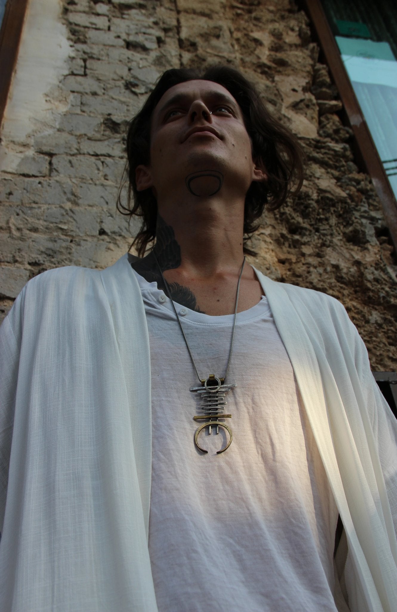 A silver and brass necklace with a geometric pendant, featuring a chain with small bell-like charms and a larger brass ring pendant.